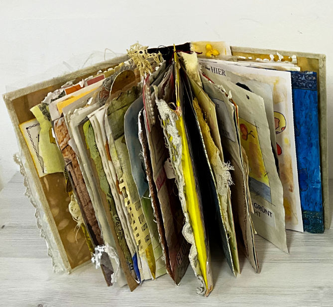 Finished Golden Threads mixed media art journal standing open on a wooden surface, thick with collage pages, lace and fabric covers, shown from the side to reveal its "gator mouth" spine.